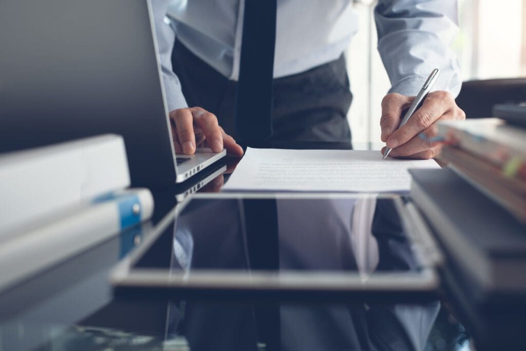 A person in professional attire stands at a desk, writing on a lease document. A laptop, tablet, and various office supplies are visible on the desk, suggesting a work or business environment. The scene is well-lit.